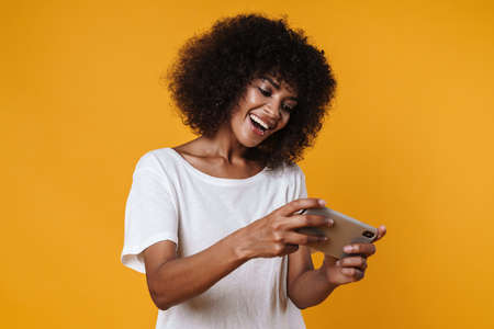 Image of excited african american girl playing video game on mobile phone isolated over yellow wallの写真素材