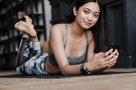 Image of cheerful asian girl in sportswear using mobile phone while lying on yoga mat at home