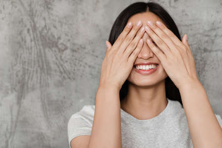 Image of joyful beautiful asian girl laughing and covering her eyes while posing indoorsの写真素材