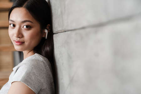 Image of beautiful asian girl using earphones while leaning on concrete wall indoorsの写真素材