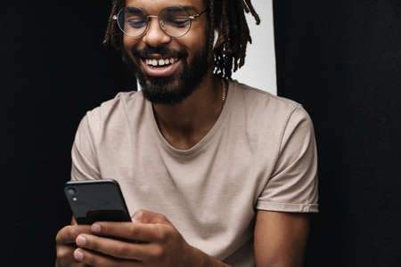 Photo of african happy laughing guy isolated over black and white wall background using mobile phone...
