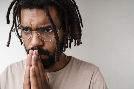 Photo of handsome serious praying african guy isolated over grey wall background in eyeglasses