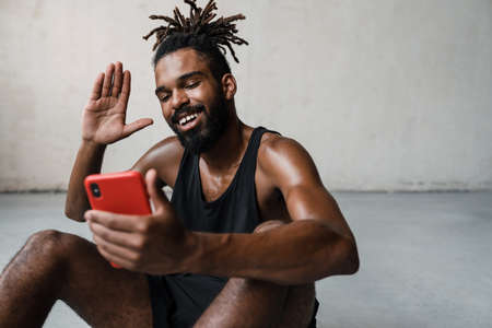 Image of smiling african american sportsman waving hand while using mobile phone indoorsの写真素材