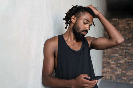 Image of serious african american sportsman using cellphone while leaning on wall indoors