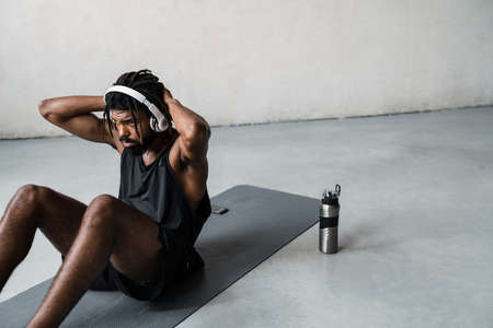 Image of strong african american sportsman in headphones doing exercise while working out on mat indoorsの写真素材