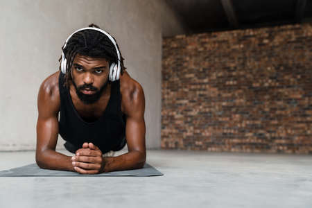 Image of strong african american sportsman in headphones doing exercise while working out on mat indoorsの写真素材