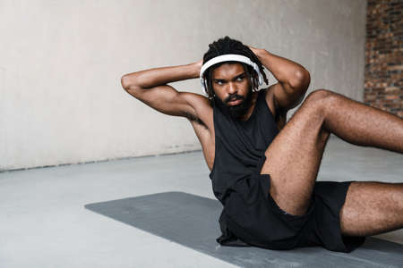Image of strong african american sportsman in headphones doing exercise while working out on mat indoorsの写真素材