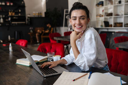 Smiling young woman studying on laptop computer while sitting in cafe indoors