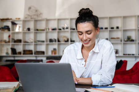 Smiling young woman studying on laptop computer while sitting in cafe indoors