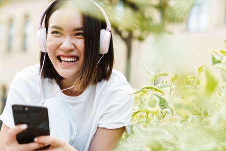 Photo of a laughing young asian woman sitting outdoors while using mobile phone and listening music...