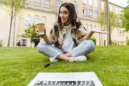Photo of pleased unsured young asian woman sitting outdoors on grass using laptop computer and...