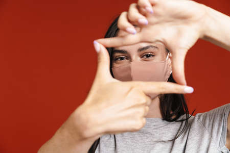 Image of young woman in face mask making photo frame gesture isolated over red backgroundの写真素材