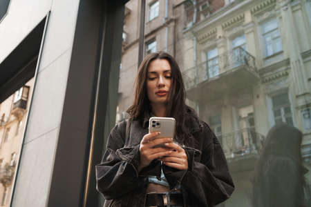 Image of concentrated young woman walking outdoors by the street and using mobile phoneの写真素材
