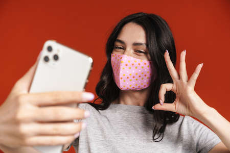 Image of young joyful woman in face mask taking selfie on mobile phone isolated over red backgroundの写真素材