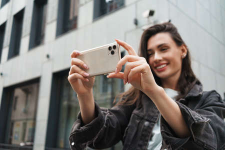 Image of positive pleased young woman walking outdoors by the street and using mobile phone