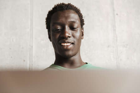 Image of optimistic young african american man in casual clothes working on laptop computer at home