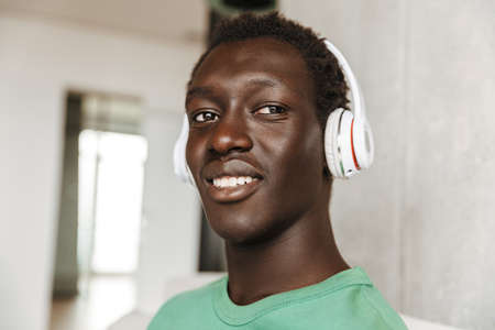 Image of satisfied joyful african american man in headphones sitting on sofa at home