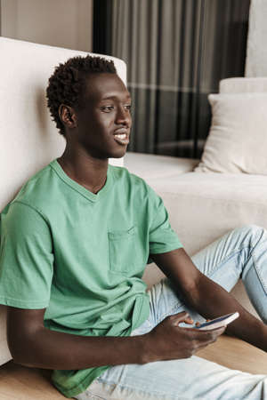 Image of attractive happy african american man in casual clothes holding smartphone while sitting on floor at homeの写真素材