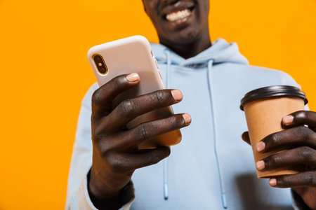 Image of young african american guy in hoodie holding cellphone and takeway coffee cup isolated over...