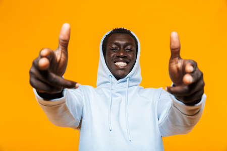 Image of pleased african american guy in streetwear hoodie smiling and pointing fingers at you isolated over yellow backgroundの写真素材