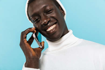 Image of happy african american guy in white clothes smiling and talking on cellphone isolated over...
