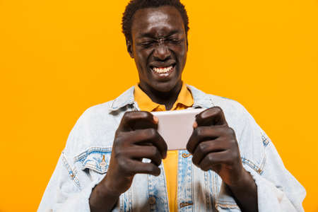 Image of young african american guy in denim jacket smiling and holding cellphone isolated over...