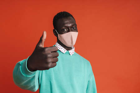 Confident african man wearing protective medical mask showing thumbs up isolated over red background