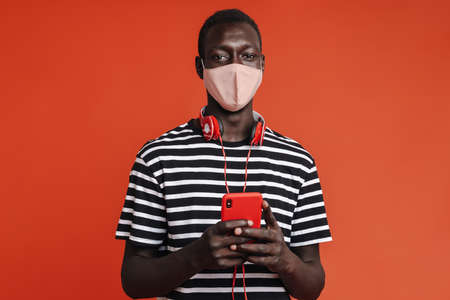 Afro american man with medical mask holding cellphone isolated on red background