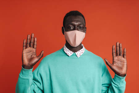 Serious afro american man wearing medical mask showing stop gesture isolated over red background