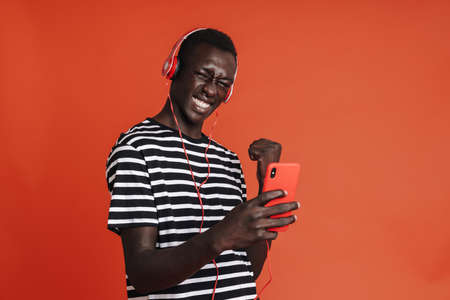 Smiling young afro american man in headphones using mobile phone while standing isolated over red...