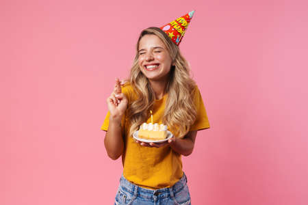 Photo of dreaming birthday woman isolated over pink wall background holding cake with candle and...