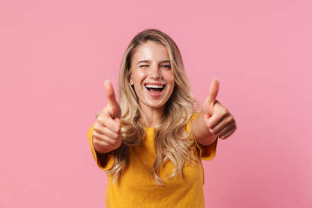 Image of a smiling winking young woman posing isolated over pink wall background showing thumbs up...