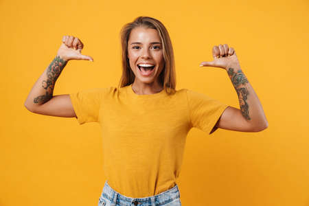 Happy young girl pointing fingers at blank t-shirt isolated over yellow background