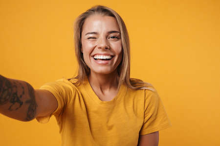 Image of young joyful woman winking and taking selfie photo isolated over yellow background