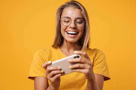 Image of smiling woman in eyeglasses playing online game on cellphone isolated over yellow backgroundの写真素材