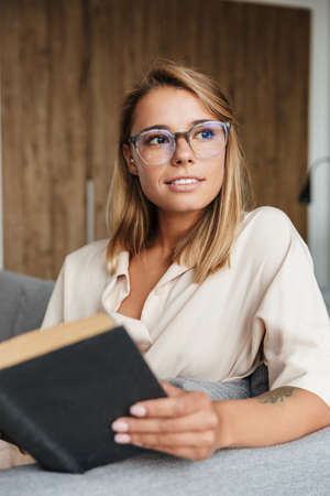 Image of young pleased woman in pajama reading book while sitting on couch at home