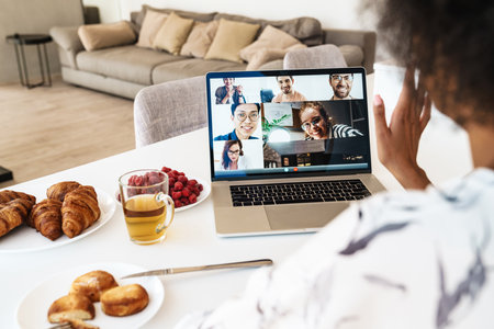 Cheerful beautiful woman using laptop for a video call while having breakfast in modern kitchen,...