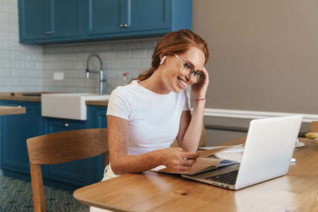 Image of smiling ginger woman in earphones working with laptop while sitting at home kitchen