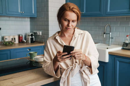 Image of serious redhead woman using cellphone while standing at home kitchenの写真素材