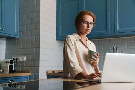 Image of thinking ginger woman drinking coffee and using laptop while sitting at home kitchenの写真素材
