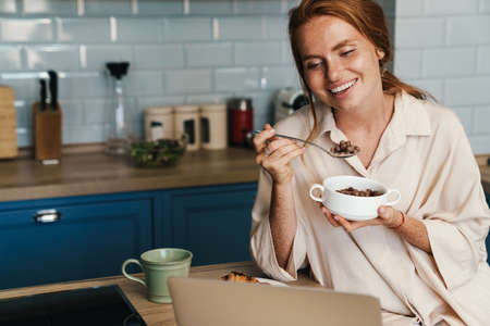 Image of cheerful ginger woman using laptop while having breakfast at home kitchen