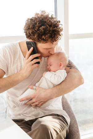 Attractive young father talking on mobile phone while sitting at the table at home and holding his...