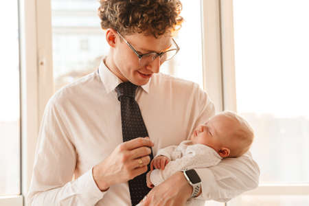 Young smiling father businessman playing with his little baby son while standing at the window at...