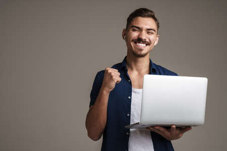 Image of happy handsome guy showing winner gesture while holding laptop isolated over grey backgroundの写真素材