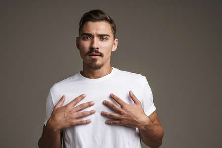 Image of confused unshaven guy posing with hands on his chest isolated over grey background