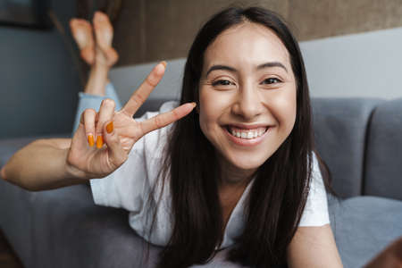 Image of a smiling young woman at home indoors lying on a sofa and taking a selfie by camera with...