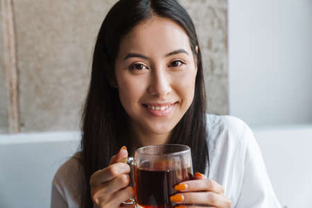 Image of a smiling young woman sitting at home indoors and drinking tea