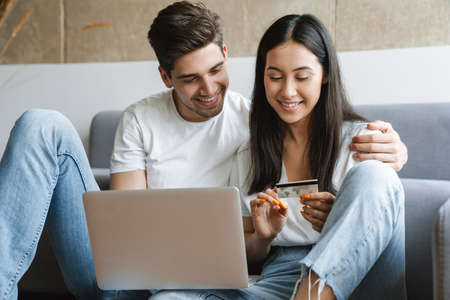 Photo of a cheerful young loving couple at home on floor near sofa using laptop computer and holding credit cardの写真素材