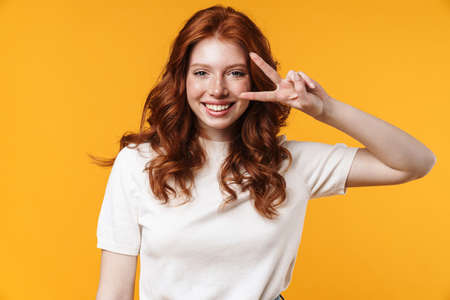 Image of ginger joyful girl winking and gesturing peace sign isolated over yellow backgroundの写真素材