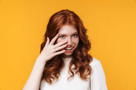 Image of ginger joyful girl smiling and covering er face isolated over yellow backgroundの写真素材